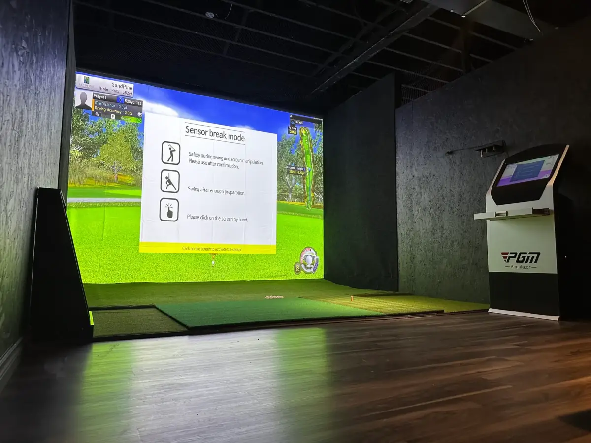 An empty simulator bay at PGM Indoor Golf Calgary with the hitting mat, projection screen and touchscreen beside it