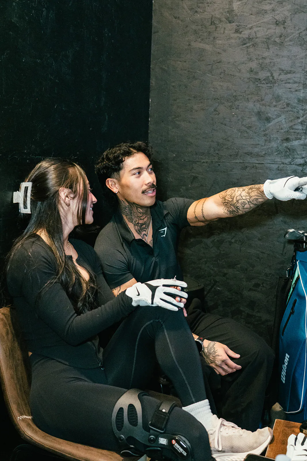 Two golfers sit on the bay chairs while one points at the simulator screen