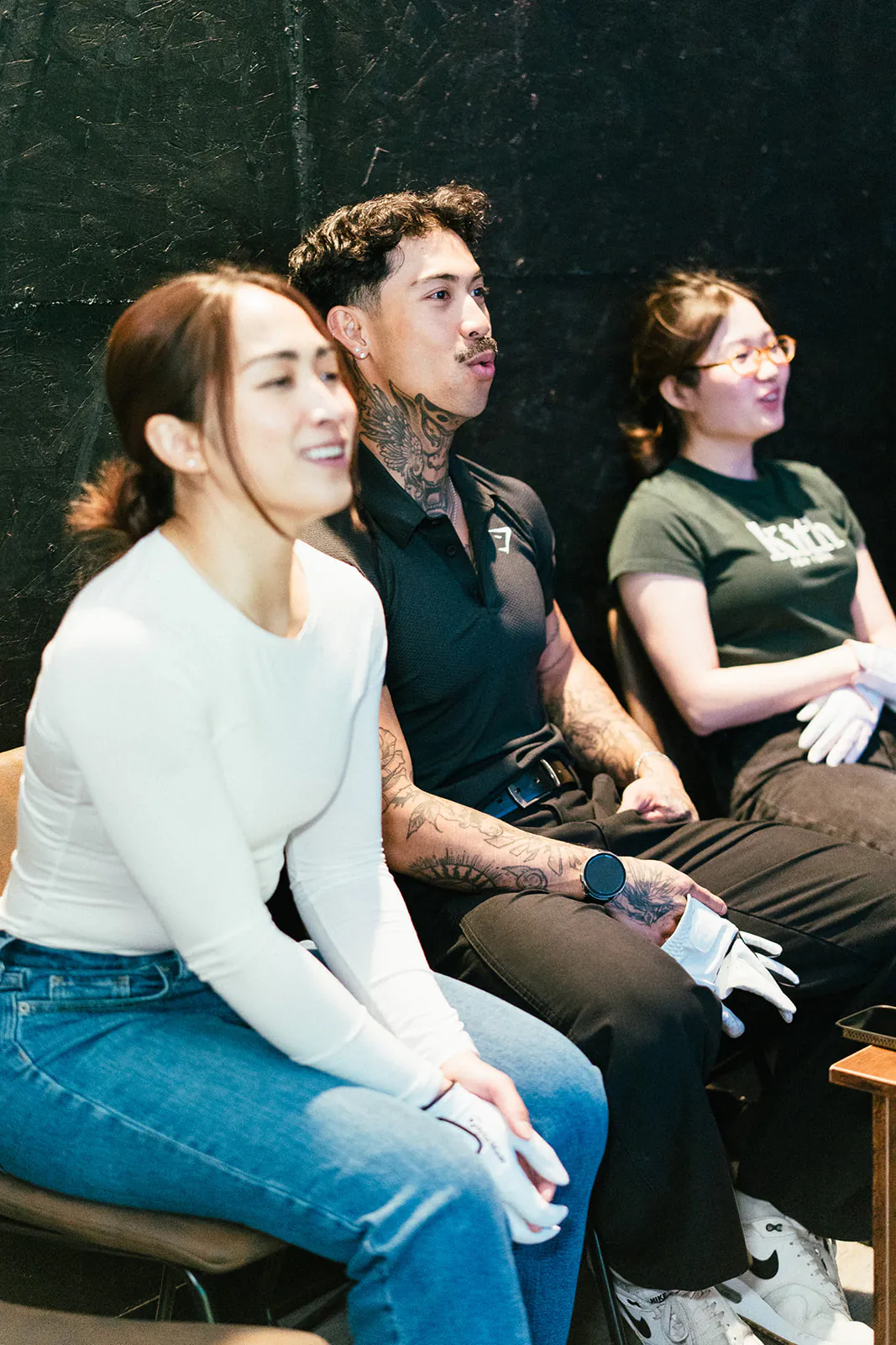 Three colleagues sit on the chairs beside the hitting mat, watching a shot