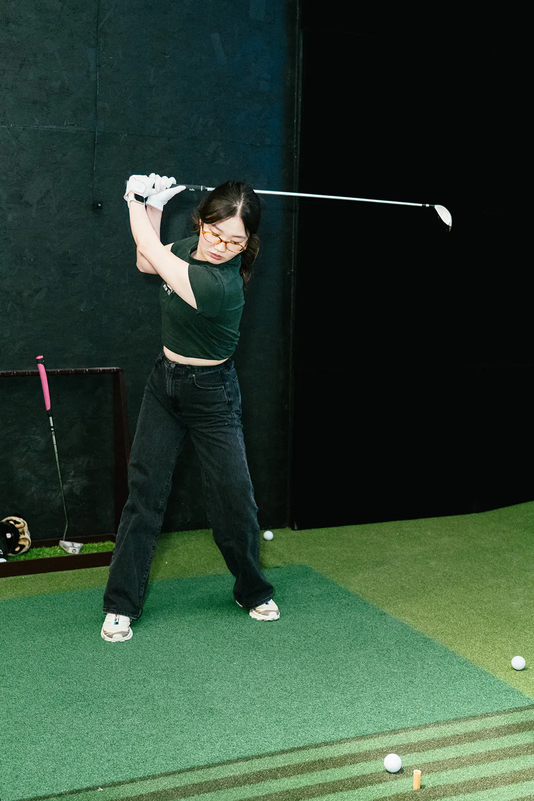 A golfer at the top of her backswing on the hitting mat in a dark PGM simulator bay