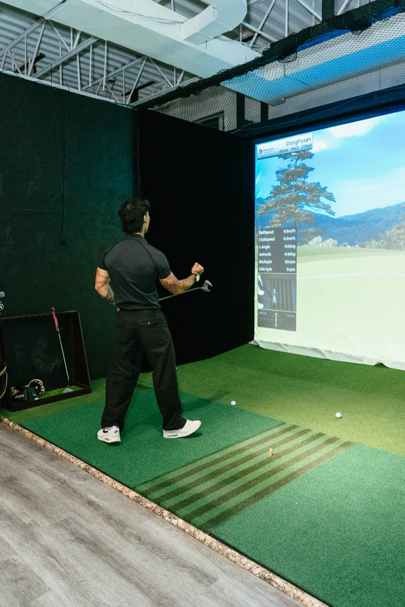 A golfer watches his shot fly, with ball data and a mountain course on the simulator screen