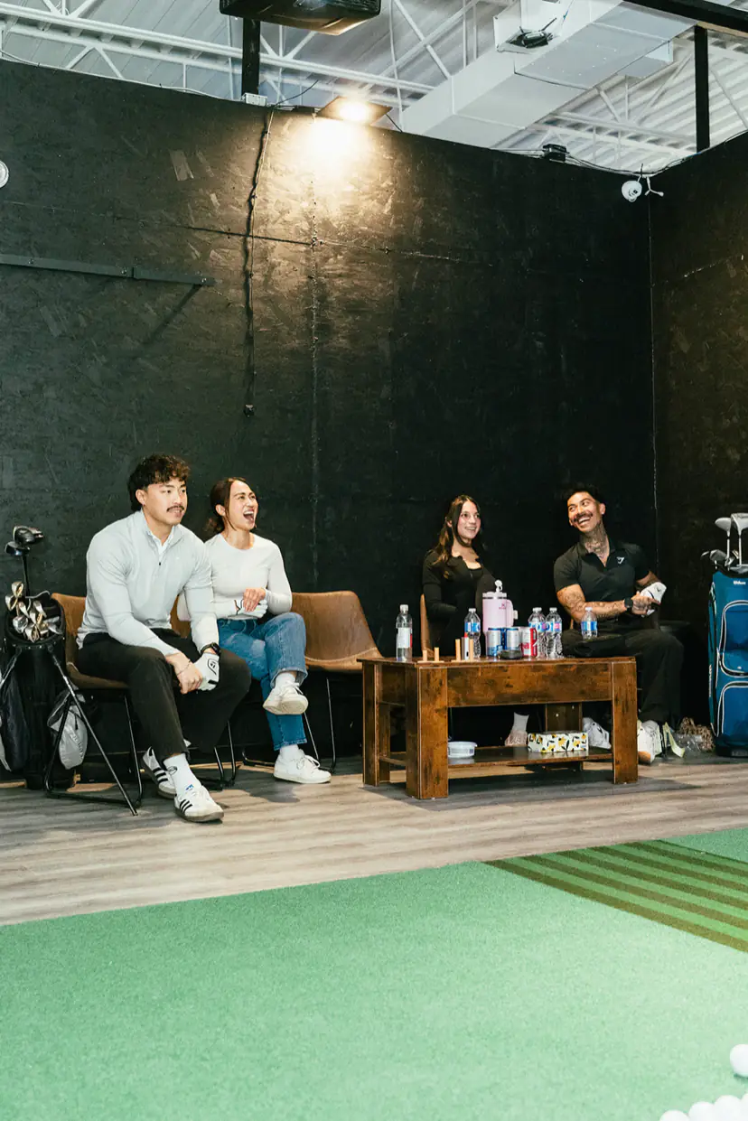 Four friends watch from the chairs beside the hitting mat in a PGM simulator bay