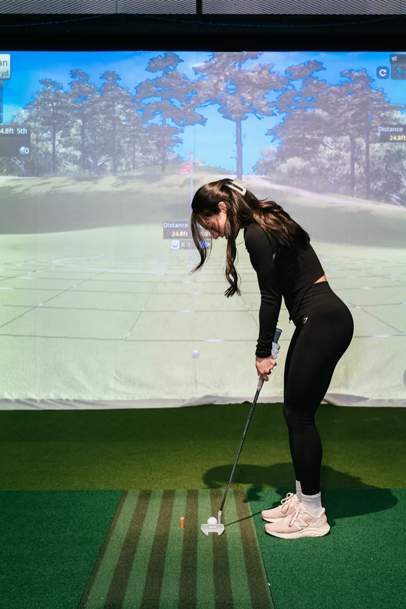 A golfer lines up a putt on the simulator green, with the distance to the hole on screen