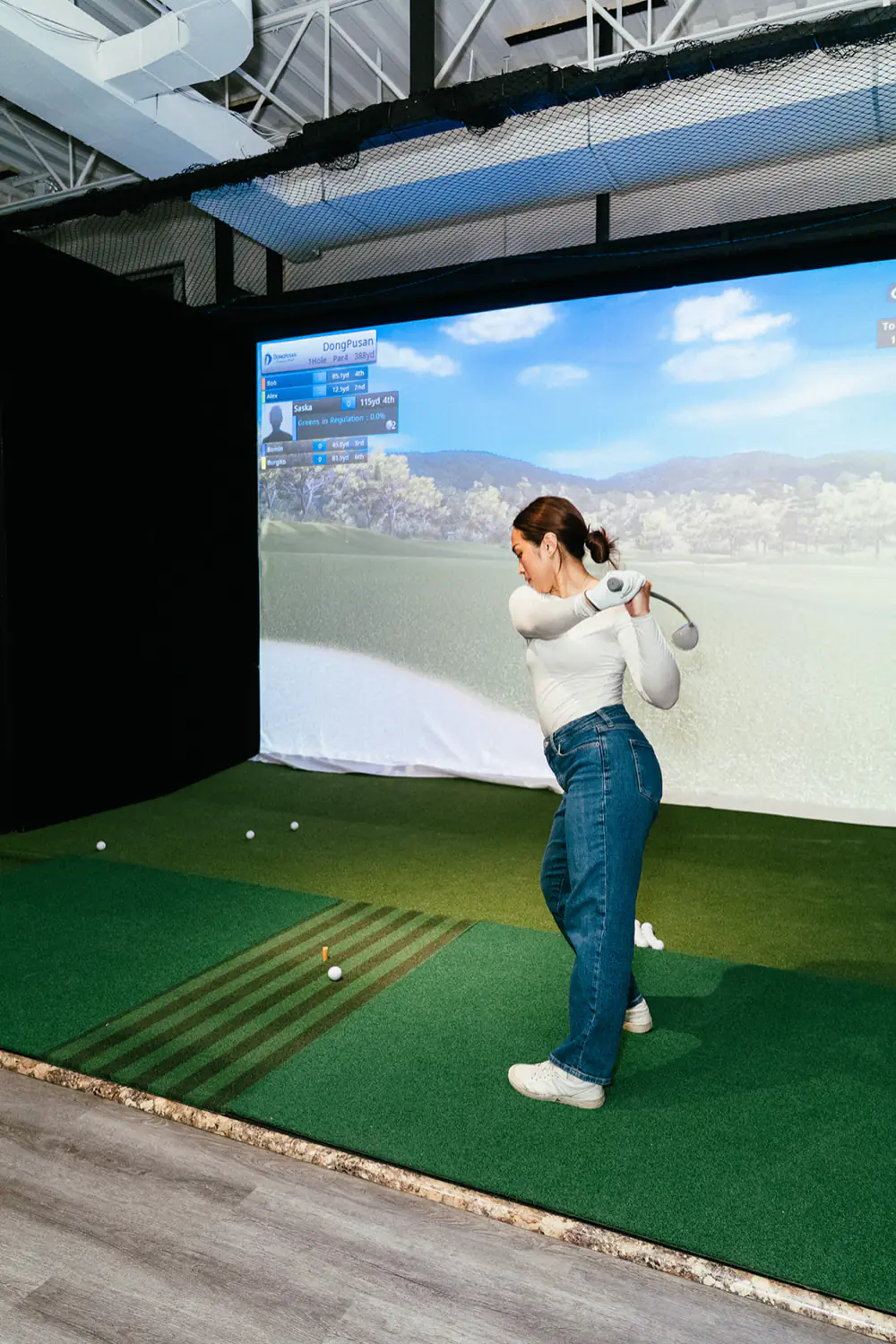 A golfer at the top of her backswing in a private PGM simulator bay, facing a projected course