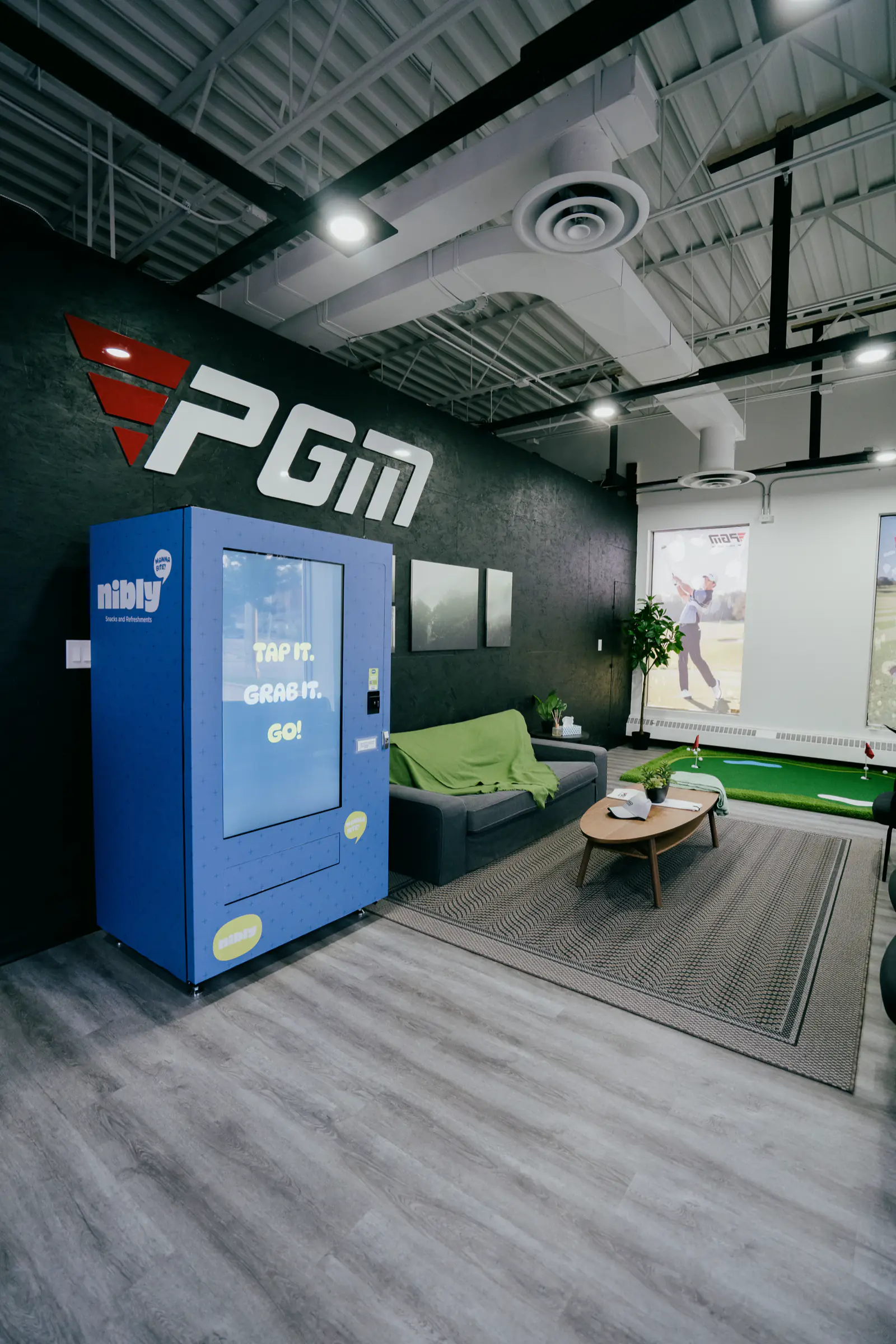 The PGM lounge with a sofa, putting green and a snack and drink vending machine under the logo wall