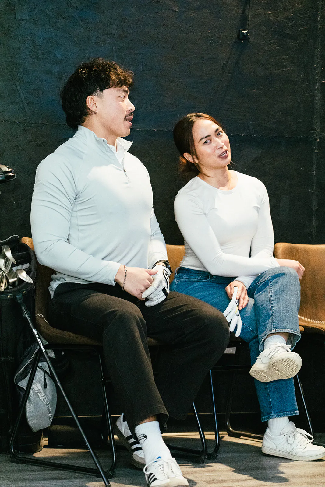 Two friends, a man and a woman, sit on chairs in a dark simulator bay reacting to a shot, with their clubs beside them