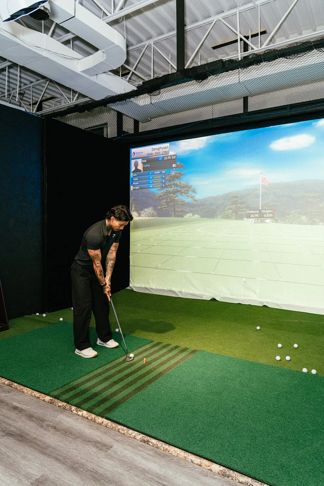 A golfer sets up to a ball on the hitting mat in front of a large course screen showing a fairway lined with trees