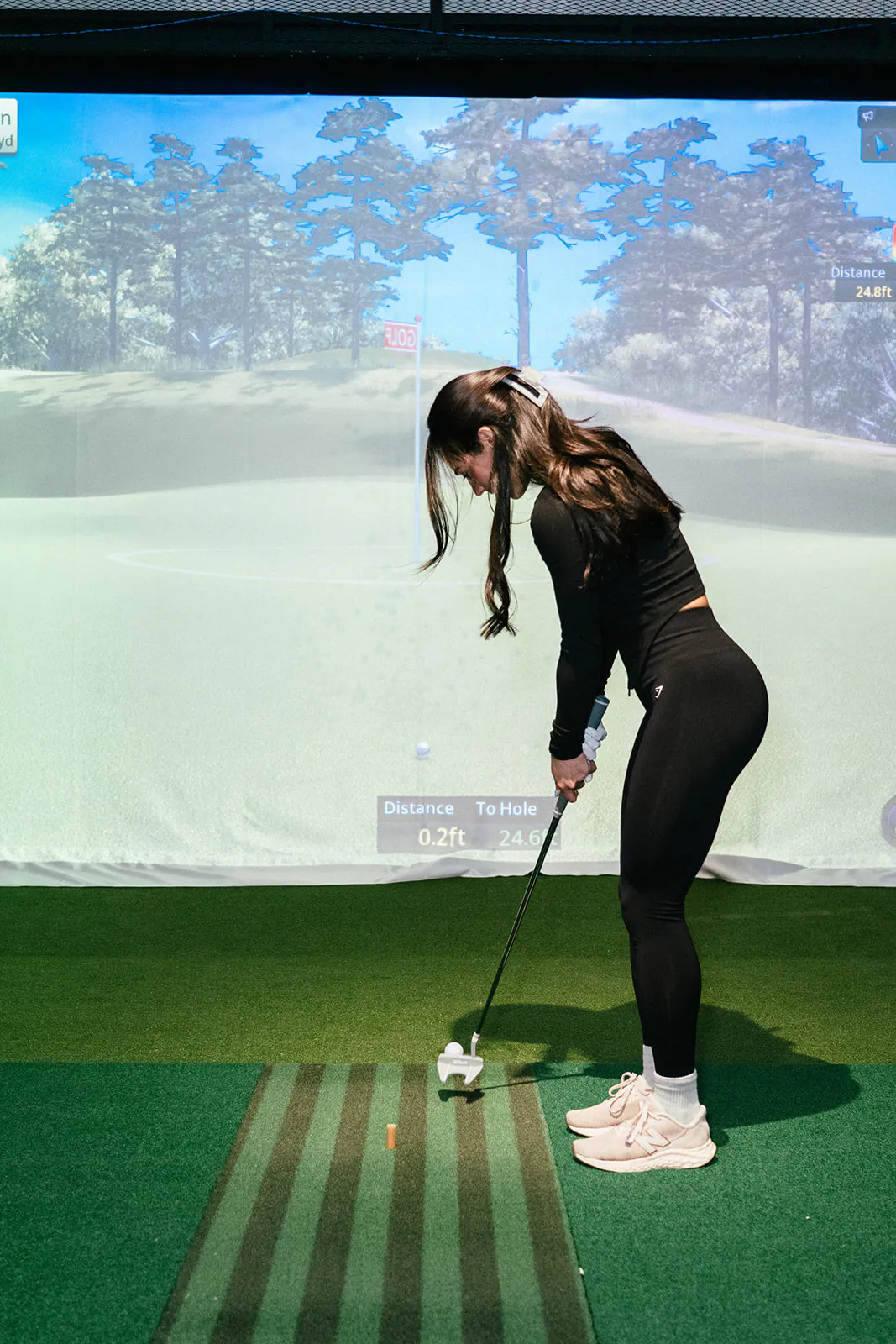 A golfer addresses the ball on the hitting mat with a course scene and shot data on the simulator screen