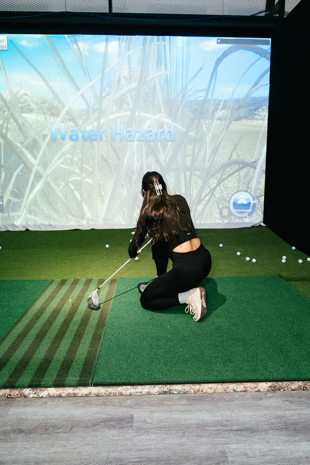 A golfer kneels on the hitting mat to tee up a ball in front of the course screen