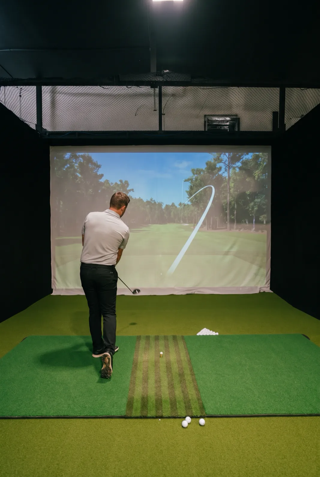 A golfer seen from behind in a simulator bay, with a ball flight on the screen that curves to the right