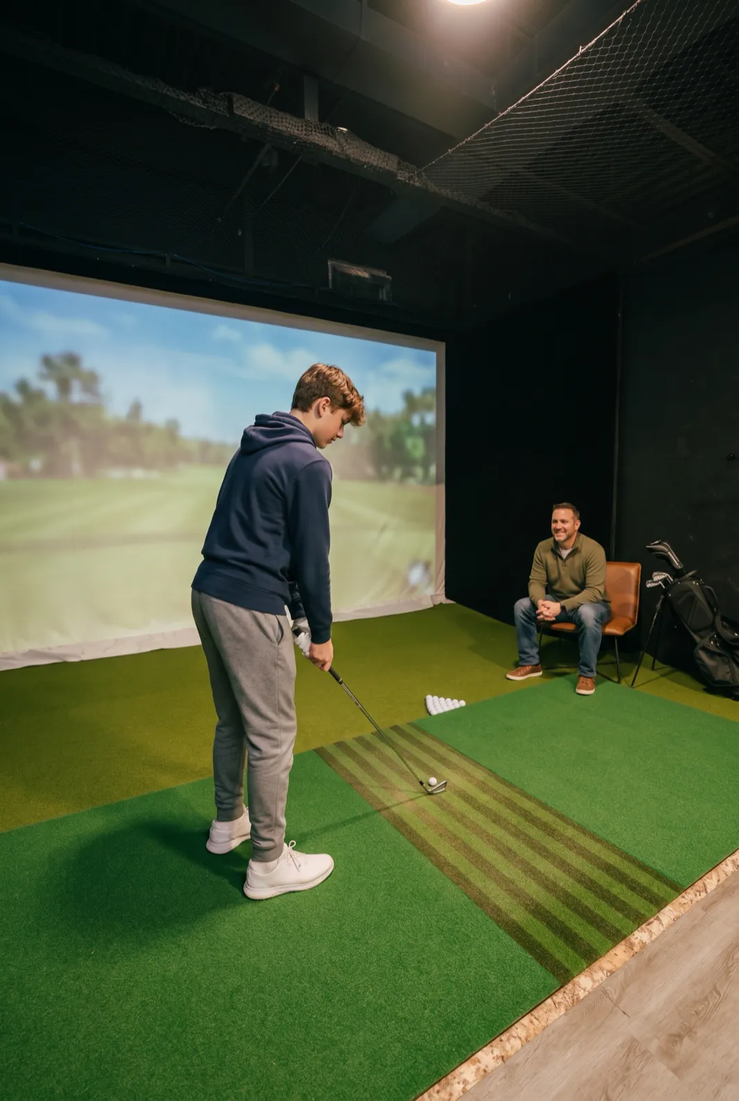 A teenager sets up to hit an iron in a simulator bay while his father watches from a chair behind him