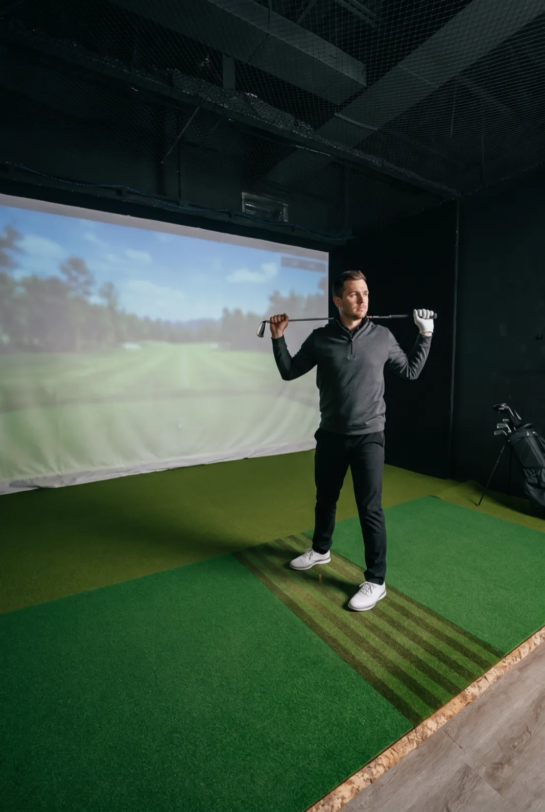 A golfer in a simulator bay holds an iron across his shoulders and turns his upper body to warm up