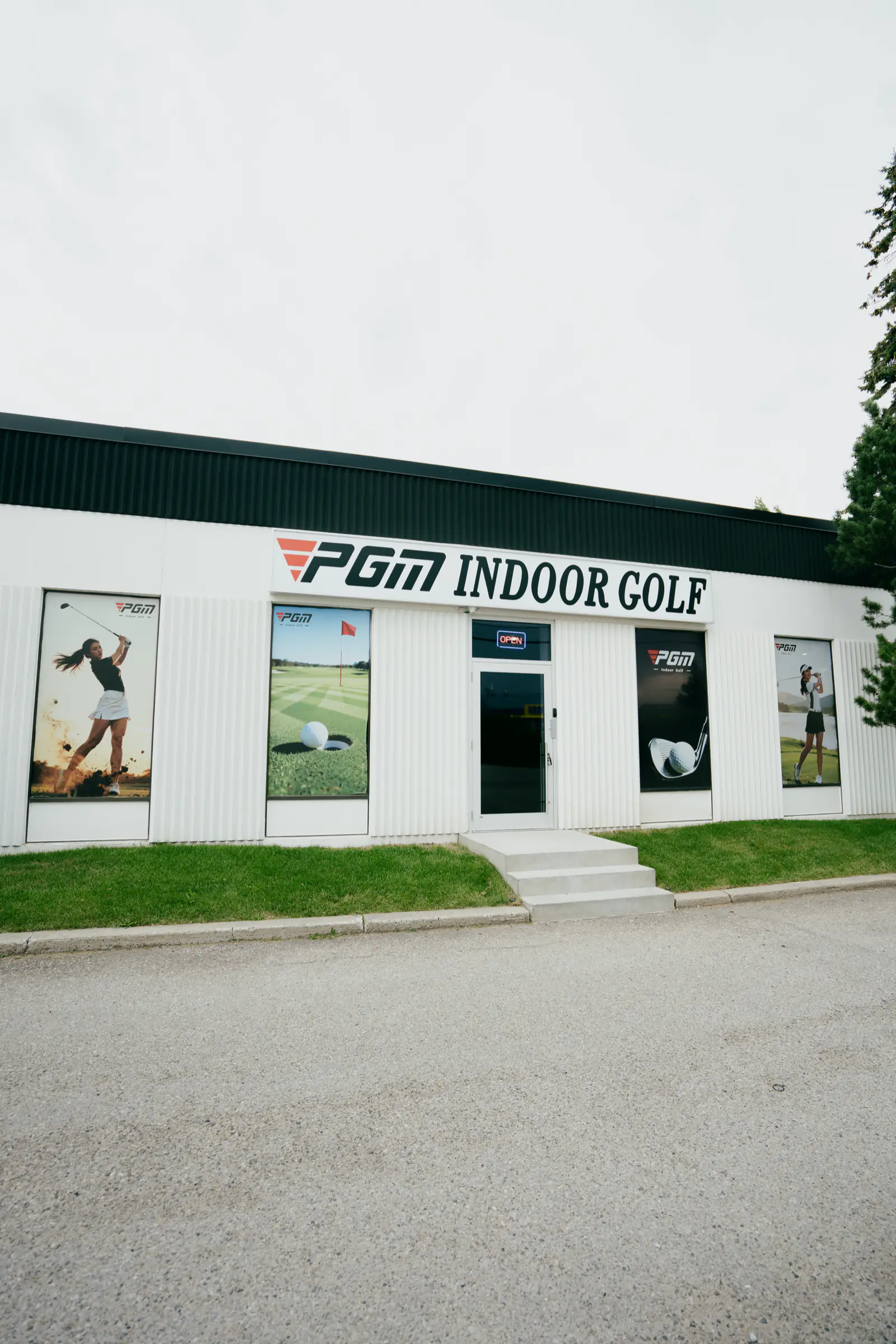 The PGM Indoor Golf storefront seen from the parking lot, a white building with the PGM Indoor Golf sign and golfer graphics in the windows